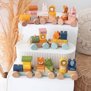 Peut inclure: Trois trains jouets en bois avec des blocs aux tons pastel et terre. Chaque train affiche des prénoms, dates de naissance, heures, poids en grammes et tailles en centimètres. Les blocs incluent des figurines d'animaux comme des lions et renards.