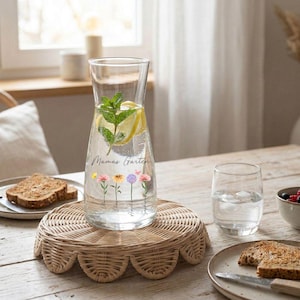 May include: A clear glass water pitcher filled with water, lemon slices, and mint leaves. The pitcher has the words "Mamas Garten" and floral illustrations. It sits on a round, woven wicker tray. A glass of water and a plate of toast are nearby.