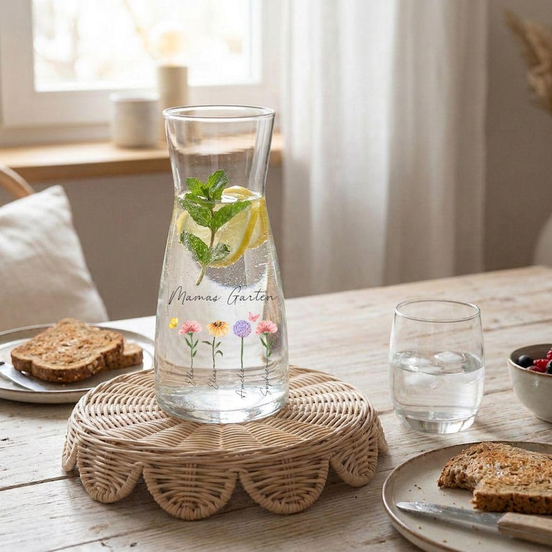 May include: A clear glass water pitcher filled with water, lemon slices, and mint leaves. The pitcher has the words "Mamas Garten" and floral illustrations. It sits on a round, woven wicker tray. A glass of water and a plate of toast are nearby.