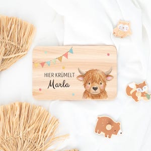 May include: A rectangular wooden plaque featuring a printed illustration of a Highland cow, colorful bunting flags, and the text "HIER KR&Uuml;MELT Marla". The plaque is accompanied by three small wooden animal-shaped cutouts of an owl, a fox, and a bear.
