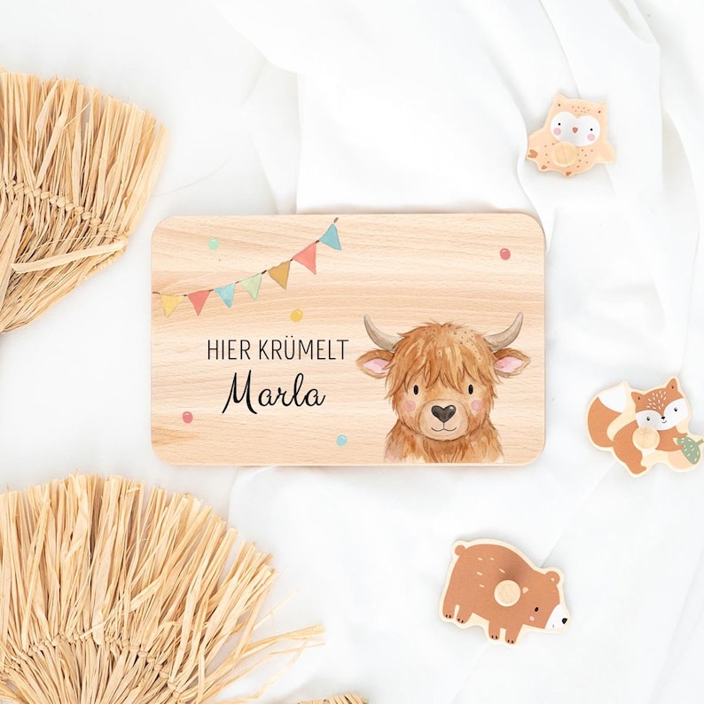 May include: A rectangular wooden plaque featuring a printed illustration of a Highland cow, colorful bunting flags, and the text "HIER KR&Uuml;MELT Marla". The plaque is accompanied by three small wooden animal-shaped cutouts of an owl, a fox, and a bear.