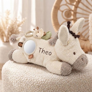 May include: A cream-coloured plush donkey toy featuring the name Theo embroidered in dark brown thread on its side. The toy includes a circular mirror, a small bird-shaped attachment, and a green leaf accent, with dark brown tufted hair.