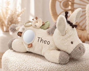 May include: A cream-colored plush donkey toy featuring the name Theo embroidered in dark brown thread on its side. The toy includes a circular mirror, a small bird-shaped attachment, and a green leaf accent, with dark brown tufted hair.