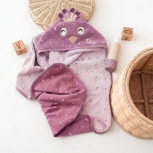 May include: A hooded baby towel in shades of purple, featuring a bird-like design on the hood with the name "Leni". The towel is accompanied by a matching washcloth, a lotion bottle, and wooden blocks. The towel has a dotted pattern.