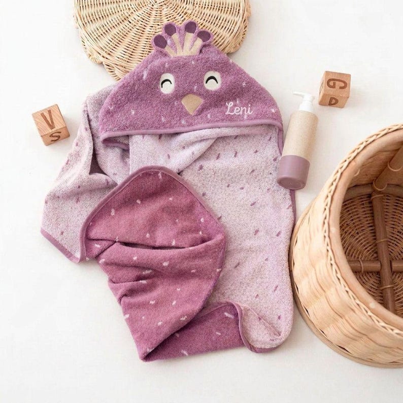 May include: A hooded baby towel in shades of purple, featuring a bird-like design on the hood with the name "Leni". The towel is accompanied by a matching washcloth, a lotion bottle, and wooden blocks. The towel has a dotted pattern.