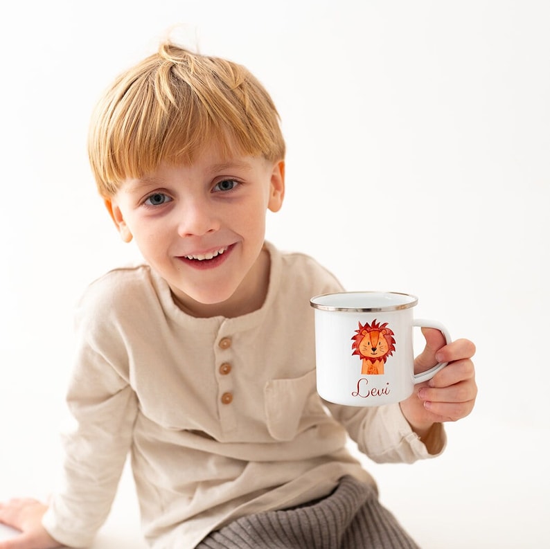 Puede incluir: Una taza de esmalte blanco con borde plateado que presenta una ilustraci&oacute;n estilo acuarela de un le&oacute;n y el nombre Levi impreso en letra roja. La taza tiene un asa en el lado derecho y se muestra sobre un fondo blanco liso.