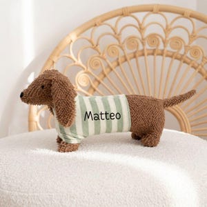 Puede incluir: Un juguete de peluche marrón con forma de perro salchicha que lleva una camiseta a rayas verdes y blancas con el nombre "Matteo" impreso. El juguete está sobre una superficie blanca, con una silla de mimbre en el fondo.