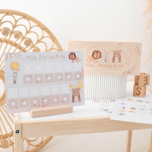 Op de afbeelding: Een houten routinekaart en kaartenset voor kinderen. De kaart toont een illustratie van een konijn, zon, beer en maan. De houten doos heeft een leeuw, konijn en beer ontwerp. Inclusief houten blokken met letters.