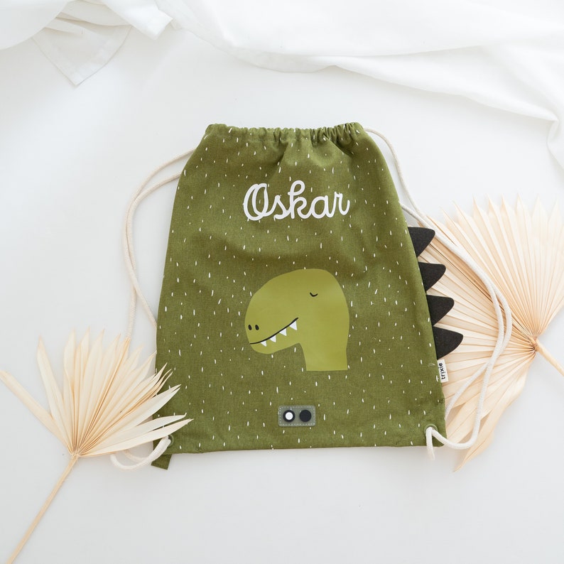 May include: Green drawstring bag with a dinosaur graphic and the name "Oskar" embroidered on the front. The bag has a white drawstring and a black and white tag.