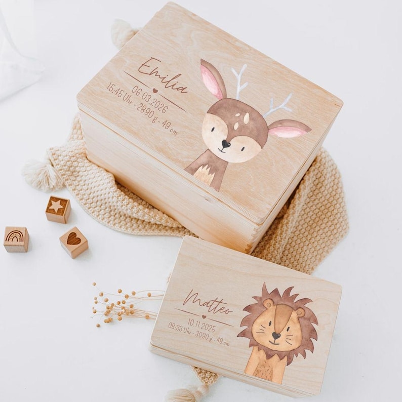 Puede incluir: Dos cajas de madera rectangulares con ilustraciones impresas de un ciervo y un le&oacute;n. La caja m&aacute;s grande muestra el nombre Emilia, una fecha y detalles de nacimiento. La caja m&aacute;s peque&ntilde;a muestra el nombre Matteo, una fecha y detalles de nacimiento.