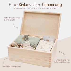 Puede incluir: Una caja de recuerdos de madera clara con tapa abatible que contiene una manta de punto verde salvia, un chupete beige, un peque&ntilde;o oso de peluche, calcetines blancos y una prenda de beb&eacute; estampada. Incluye texto en alem&aacute;n y flechas indicativas.