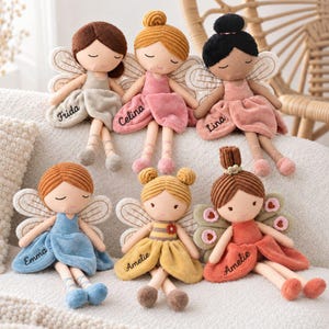 Op de afbeelding: Zes pluche poppen in feeënstijl met geborduurde namen op hun jurkjes. De poppen hebben verschillende haarkleuren, waaronder bruin, blond en zwart, in knotjes of bobs. Elke pop heeft stoffen vleugels en draagt een zachte jurk in beige, roze, blauw, geel of oranje.
