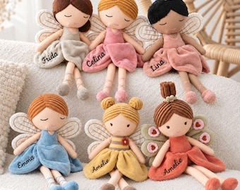 May include: Six plush fairy dolls with embroidered names on their dresses. The dolls feature various hair colors, including brown, blonde, and black, styled in buns or bobs. Each doll has fabric wings and wears a soft-textured dress in colors like beige, pink, blue, yellow, and orange.