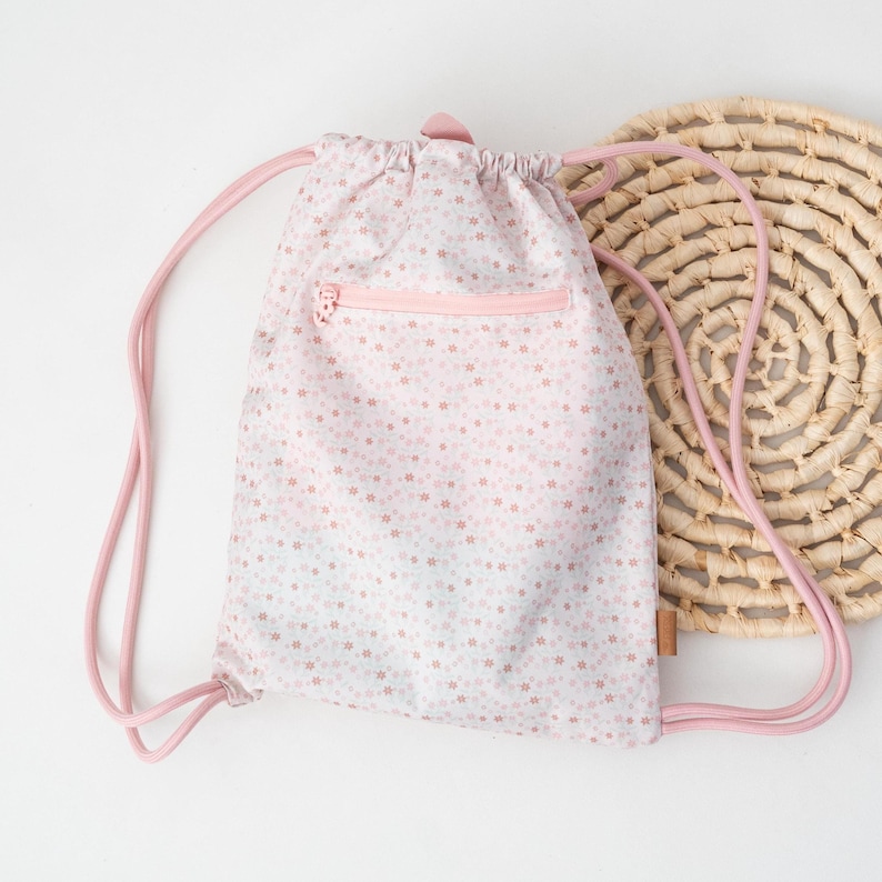 May include: A light pink drawstring backpack with a floral pattern. The bag has a pink zipper pocket and pink drawstring straps. A woven circular placemat is in the background.