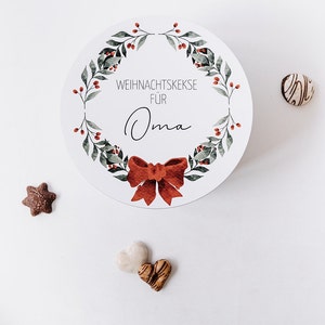 May include: A circular white container featuring a watercolour wreath design, a red bow, and the text "WEIHNACHTSKEKSE F&Uuml;R Oma". Several biscuits in various shapes are scattered around the container, including a star, heart, and round biscuit.