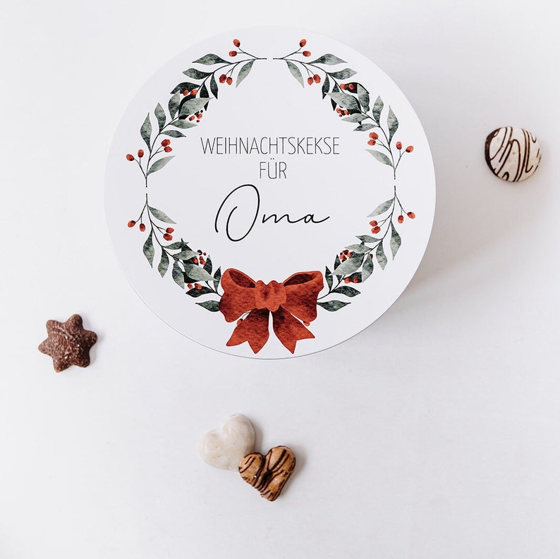 May include: A circular white container featuring a watercolour wreath design, a red bow, and the text "WEIHNACHTSKEKSE F&Uuml;R Oma". Several biscuits in various shapes are scattered around the container, including a star, heart, and round biscuit.