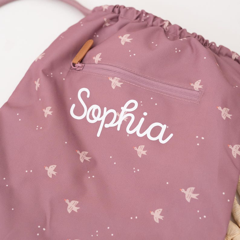 May include: A dusty rose drawstring backpack with a zipper pocket and a brown leather zipper pull. The bag is decorated with a pattern of small white birds and the name "Sophia" in white script.