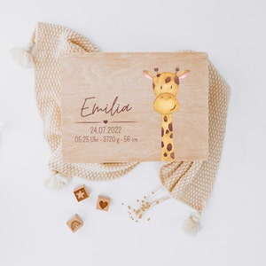Puede incluir: Una caja de recuerdos rectangular de madera con una ilustraci&oacute;n en acuarela de una jirafa a la derecha. El texto dice Emilia, 24.07.2022, y 05:25 Uhr - 3720 g - 56 cm. Tres peque&ntilde;os bloques de madera con iconos grabados de estrella, coraz&oacute;n y arco&iacute;ris.