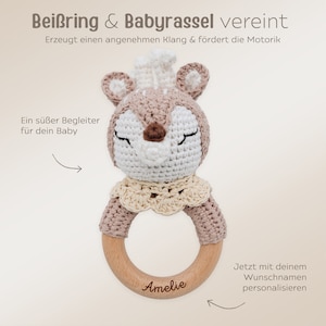 K&ouml;nnte beinhalten: Eine geh&auml;kelte Hirschratsche mit einem Holzring, personalisiert mit dem Namen "Amelie". Die Rassel ist beige und braun mit einem wei&szlig;en Gesicht. Der Text "Bei&szlig;ring & Babyrassel vereint" ist oben im Bild zu sehen.
