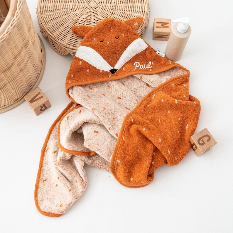 May include: An orange and white hooded baby towel shaped like a fox. The towel has a fox face design on the hood, with the name "Paul" embroidered on the side. The towel is made of soft, absorbent material, perfect for bath time.