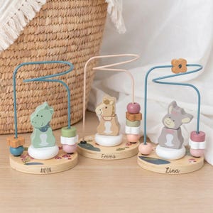 Könnte beinhalten: Drei Holz-Perlenlabyrinth-Spielzeuge mit Tierfiguren: ein grüner Dinosaurier, ein beiges Pferd und ein grauer Esel. Jedes Spielzeug hat eine Drahtbahn mit bunten Perlen und eine Holzbasis mit einem Namen. Im Hintergrund ist ein Weidenkorb.