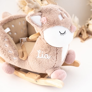 May include: A plush brown rocking deer with a white face, pink hooves, and a pink flower on its head. The deer has a safety harness and the name "Lia" embroidered on its side.