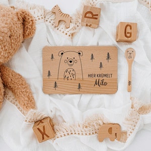 May include: A wooden plate with a bear illustration and the text "Hier kr&uuml;melt Milo". The plate is surrounded by wooden toys, including a bear, a giraffe, an elephant, and blocks with letters.
