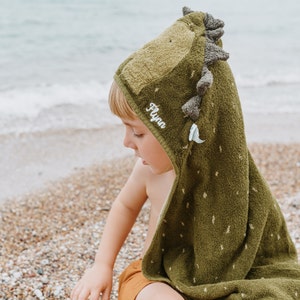 May include: A green hooded towel with a dinosaur design. The towel has a hood with a dinosaur head, complete with spikes and a small white tooth. The name "Flynn" is embroidered on the hood. The towel is on a beach.