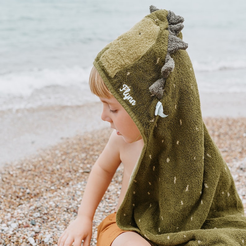 May include: A green hooded towel with a dinosaur design. The towel has a hood with a dinosaur head, complete with spikes and a small white tooth. The name "Flynn" is embroidered on the hood. The towel is on a beach.