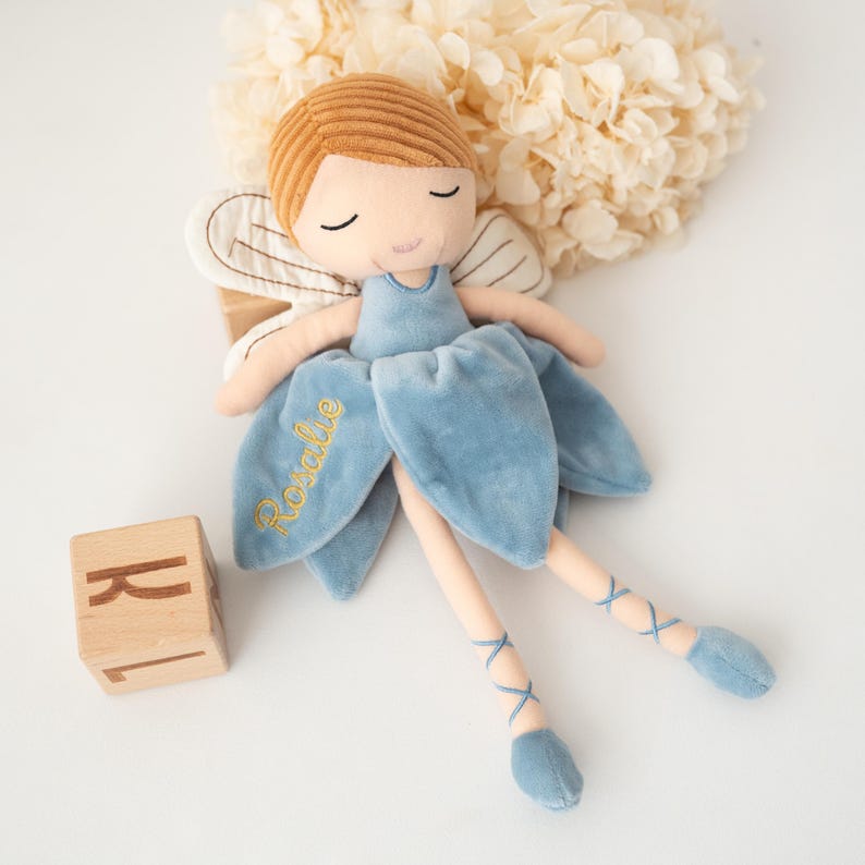 May include: A plush fairy doll with a light blue dress and ballet-style legs. The doll has brown hair and closed eyes. The name "Rosalie" is embroidered in gold on the dress. A wooden block with the letter "R" is in the image.