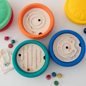 Pode incluir: Um conjunto de tabuleiros de labirinto circulares de madeira com molduras texturizadas em laranja, azul e azul-petróleo. Os tabuleiros apresentam caminhos esculpidos em espiral e lineares. Inclui pequenos piões coloridos, uma berlinde vermelha e um saco com o texto STAPEL WELT.