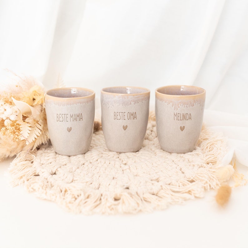 Puede incluir: Tres tazas de cer&aacute;mica beige con un s&iacute;mbolo de coraz&oacute;n y texto. Las tazas est&aacute;n inscritas con "BESTE MAMA", "BESTE OMA" y "MELINDA". Las tazas est&aacute;n sobre un posavasos de macram&eacute; color crema, con un fondo blanco y flores secas.