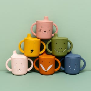 Op de afbeelding: Set van zes siliconen bekers met dierengezichten en deksels. De bekers zijn in verschillende kleuren, waaronder roze, geel, groen, oranje en blauw. Elke beker heeft twee handvatten.