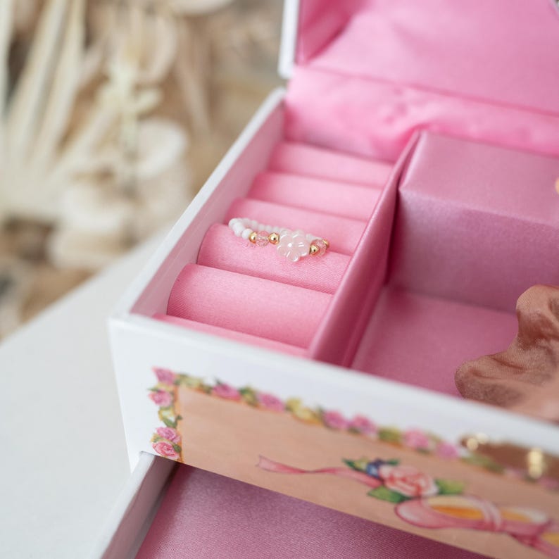 May include: A white jewelry box with pink velvet lining and a ring holder. The ring holder has a pink and white beaded ring with gold accents.