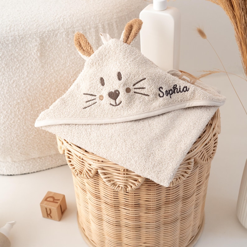 May include: A beige hooded baby towel with embroidered bunny face details and the name "Sophia". The towel is folded over a wicker basket. The bunny ears are a darker beige color.