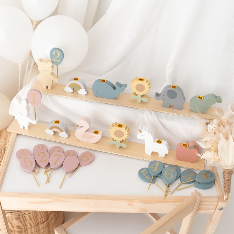 May include: Wooden animal and number decorations. The set includes a bear, rainbow, whale, sunflower, elephant, and more. Numbered balloon and circle toppers are also included. The items are displayed on a wooden stand.