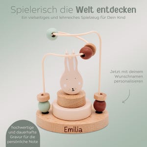 Peut inclure: Jouet en bois pour b&eacute;b&eacute; avec un lapin et des perles color&eacute;es. Le jouet a une base en bois clair avec le nom "Emilia" grav&eacute;. Le texte "Spielerisch die Welt entdecken" est en haut.