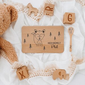 May include: A wooden tray with a fox illustration and the text "HIER KR&Uuml;MELT Phil". The tray is surrounded by wooden toys, including a giraffe, an elephant, and blocks that spell "R" and "G".