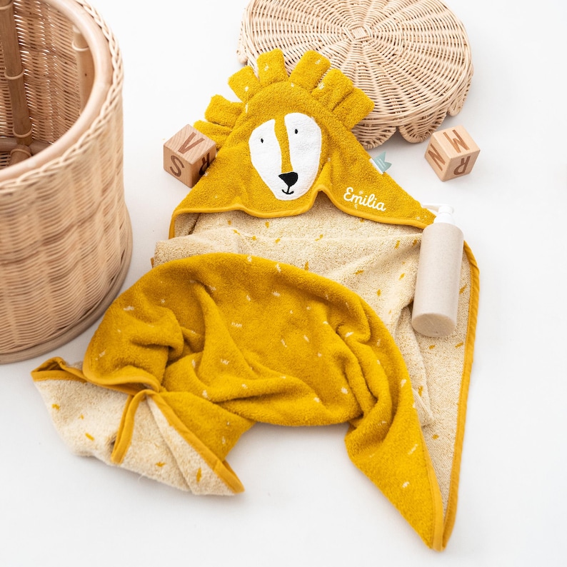 May include: A yellow hooded baby towel with a lion face design. The towel has a white face, black nose, and the name "Emilia" embroidered on it. The towel is folded and has a dotted pattern.