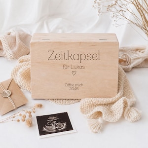 May include: A rectangular light-coloured wooden box with the engraved text Zeitkapsel für Lukas, a heart symbol, and the text Öffne mich 2046. The box is positioned with a small brown envelope, a sonogram image, and a textured cream-coloured fabric.