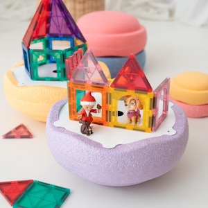 Puede incluir: Estructuras de baldosas magnéticas de colores con figuras en miniatura de un gnomo y un perro de dibujos animados. Las estructuras están sobre plataformas redondeadas de colores pastel. Hay baldosas adicionales dispersas.