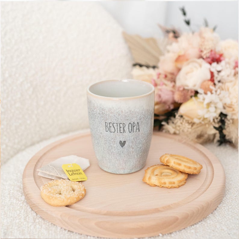 Puede incluir: Una taza de cer&aacute;mica gris con el texto "BESTER OPA" y un s&iacute;mbolo de coraz&oacute;n, colocada sobre una bandeja de madera con galletas y bolsitas de t&eacute;. La taza tiene un dise&ntilde;o moteado y un borde de color claro. Un ramo de flores est&aacute; en el fondo.