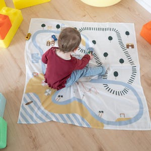 Può includere: Un bambino gioca su un tappeto bianco con un design stradale e paesaggistico. Il tappeto presenta una strada blu, un'area spiaggia gialla e alberi verdi. Piccole macchinine sono sparse sul tappeto. La parola "eano" è visibile.