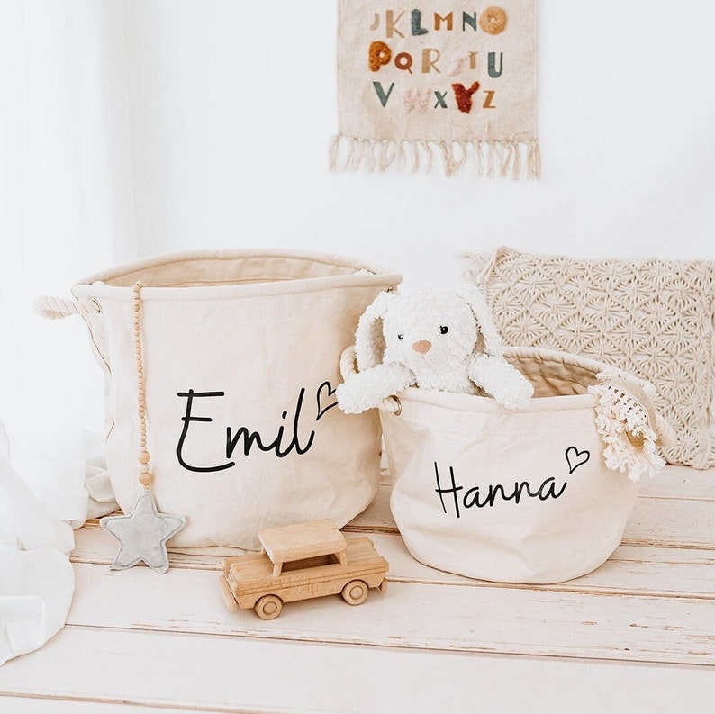 May include: Two beige canvas storage baskets with rope handles. The larger basket has the name "Emil" written on it with a heart symbol. The smaller basket has the name "Hanna" written on it with a heart symbol. A white stuffed bunny is sitting in the smaller basket.