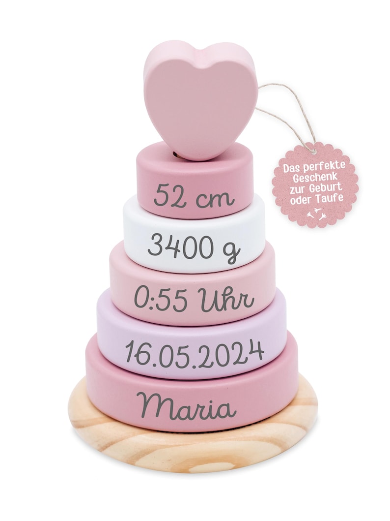 Może przedstawiać: Drewniana zabawka do układania w stosy w odcieniach r&oacute;żu, bieli i naturalnego drewna, zwieńczona sercem. Zabawka zawiera wymiary 52 cm, 3400 g i imię "Maria". Etykieta głosi: "Idealny prezent na narodziny lub chrzest."