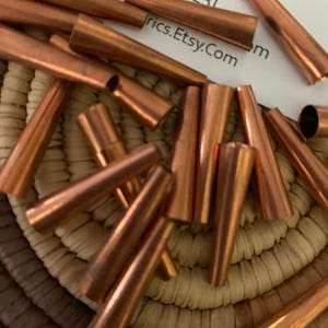 May include: A collection of copper-colored, cone-shaped metal beads. The beads are scattered on a woven basket. The image also shows a business card with the text "Bearpawfa" and "rics.Etsy.Com".