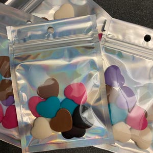 May include: A clear plastic bag filled with small heart-shaped resin pieces in various colors, including pink, brown, teal, purple, and cream.