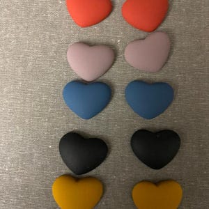 May include: A collection of ten heart-shaped objects in various colors: red, lavender, blue, black, and yellow. The hearts are arranged in pairs, with each pair of the same color. The objects are on a gray textured surface.
