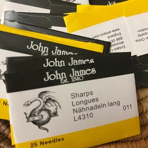 May include: A collection of yellow and black needle packets. The packets feature the "John James" logo and text indicating "Sharps" needles, with the number "L4310" and "25 Needles" visible. The packets are arranged on a woven surface.