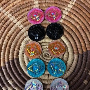 May include: Eight pairs of round earrings in various colors, including pink, black, orange, blue, and white. Each earring features a geometric design with iridescent accents. The earrings are displayed on a woven basket.
